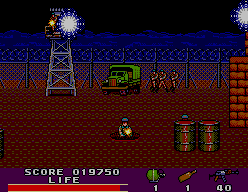 Base nocturna de Rambo III amb torres il·luminades, vehicle militar, barrils i un soldat ajupit.